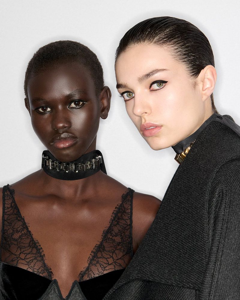 Two fashion models pose in a close-up studio shot, wearing elegant black clothing and accessories, showcasing beauty and style against a white backdrop.