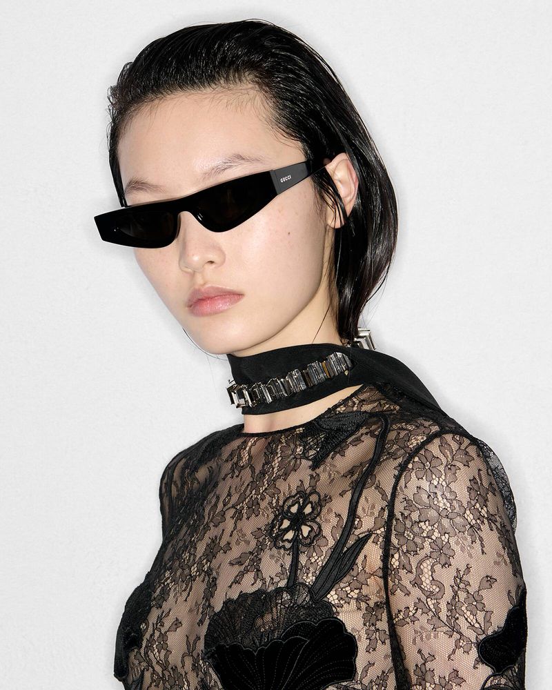 Portrait of a stylish Asian woman wearing a black lace top, black sunglasses, and a jeweled choker against a white background.
