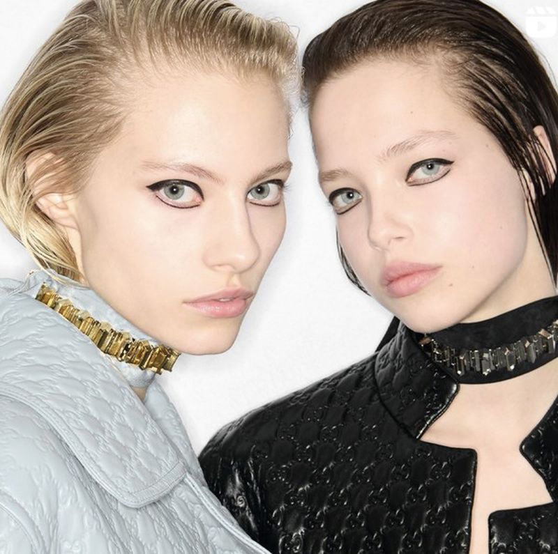 Two young women pose side-by-side with slicked-back hair, heavy eyeliner, and unique gold and silver chokers in a bright, studio setting.