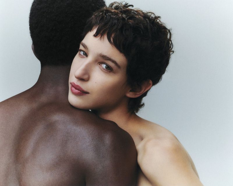 A diverse couple in a close embrace against a light backdrop, featuring a fair-skinned woman and a dark-skinned man in a studio setting.