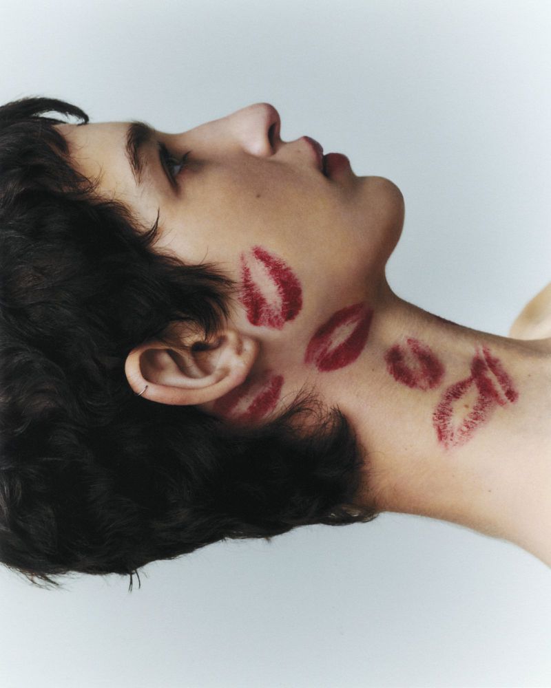 Portrait of a young adult with dark hair, looking upward, face and neck covered with dark red lipstick kiss marks against a white background.