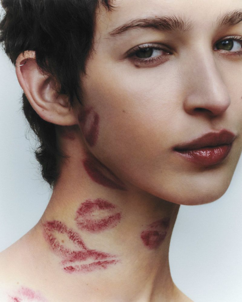 Close-up studio shot of a fair-skinned adult female with dark hair and multiple lipstick kiss marks on her face and neck.