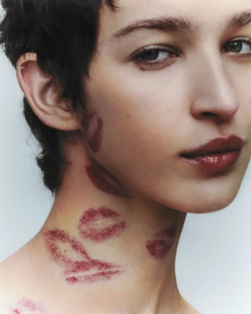 Close-up studio shot of a fair-skinned adult female with dark hair and multiple lipstick kiss marks on her face and neck.