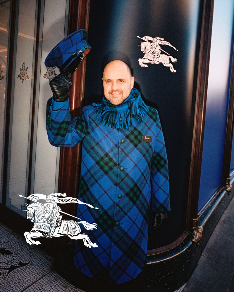 Man poses outdoors in a blue plaid coat, holding a matching hat. He is in front of a store window with equestrian logo.