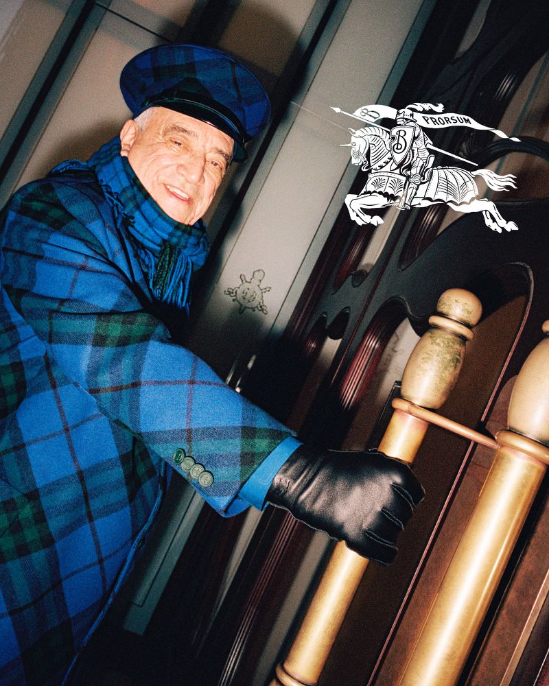 Older man in a blue plaid jacket and hat smiles at the camera. He is wearing black gloves and holding a wooden banister.