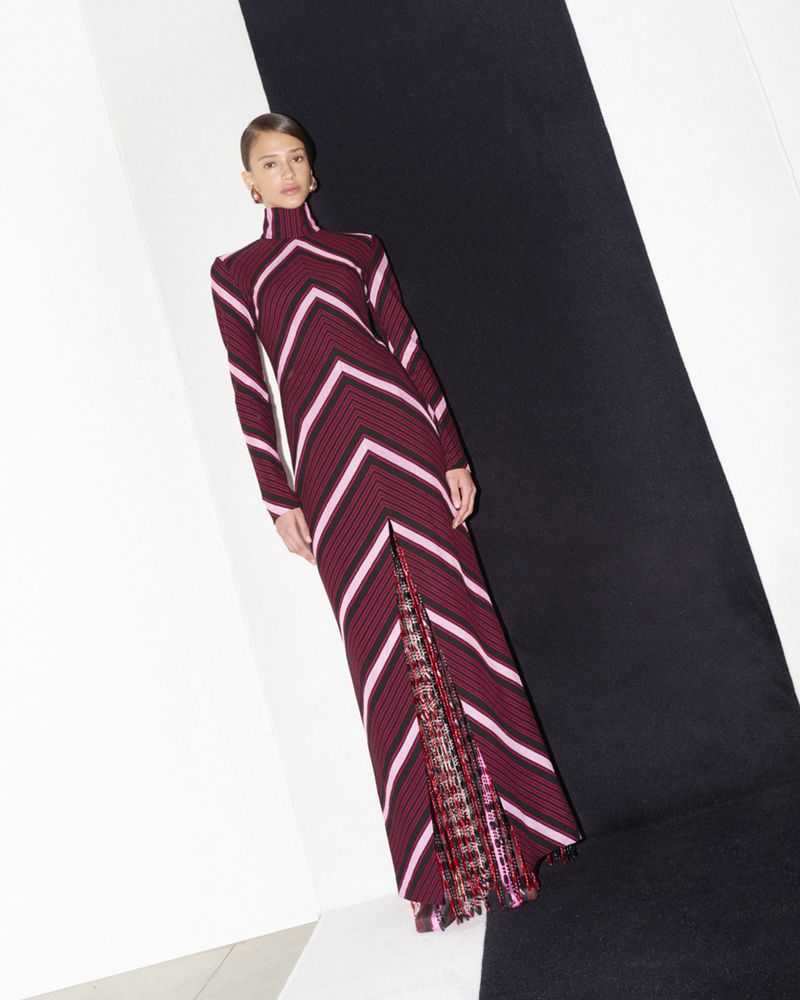 Fashion model poses in a long-sleeve, chevron-striped maroon dress with a slit, against a black and white geometric background.