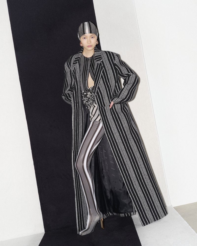 Fashion model in a striped coat, matching headscarf and leggings poses against a black and white backdrop in a studio shot.
