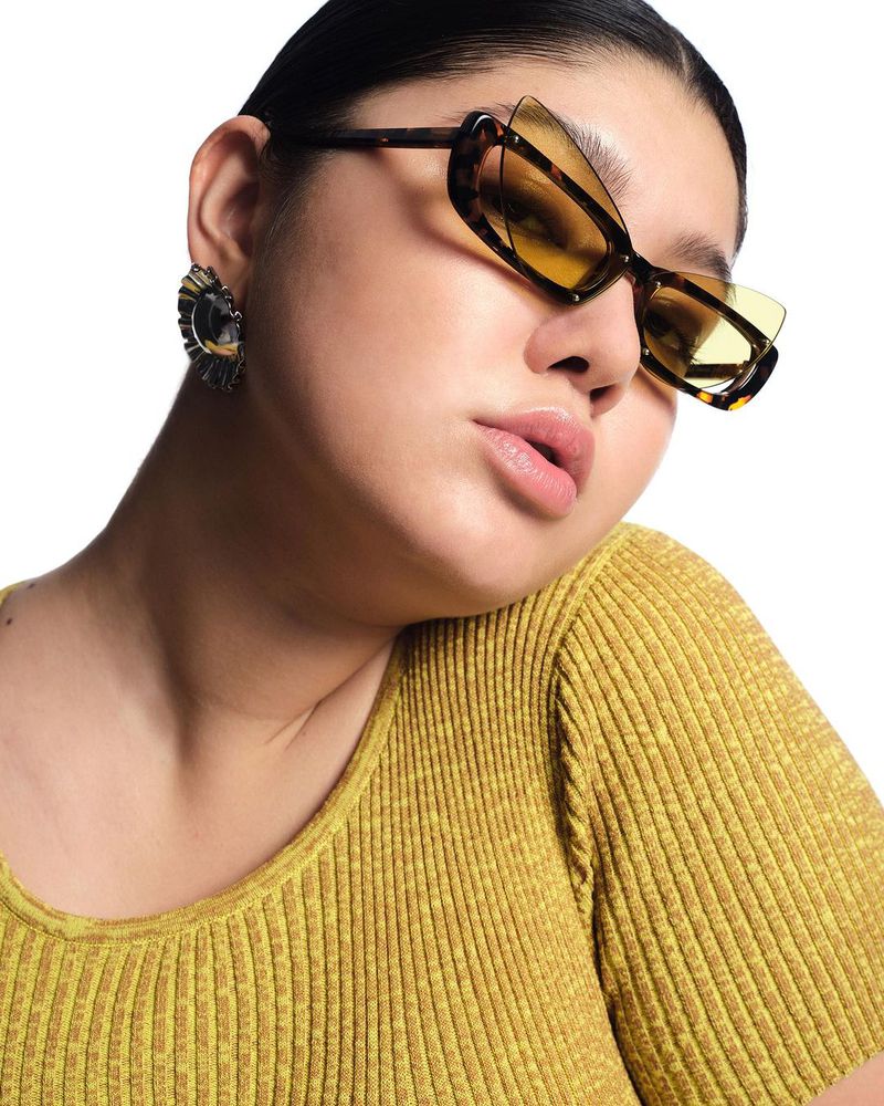 Close-up portrait of a woman wearing yellow tinted sunglasses and a yellow top, posing against a white background in a studio.