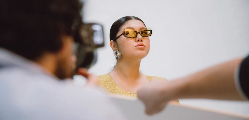 Stylish woman with yellow sunglasses and statement earrings poses in a studio with photography equipment and light modifiers visible.