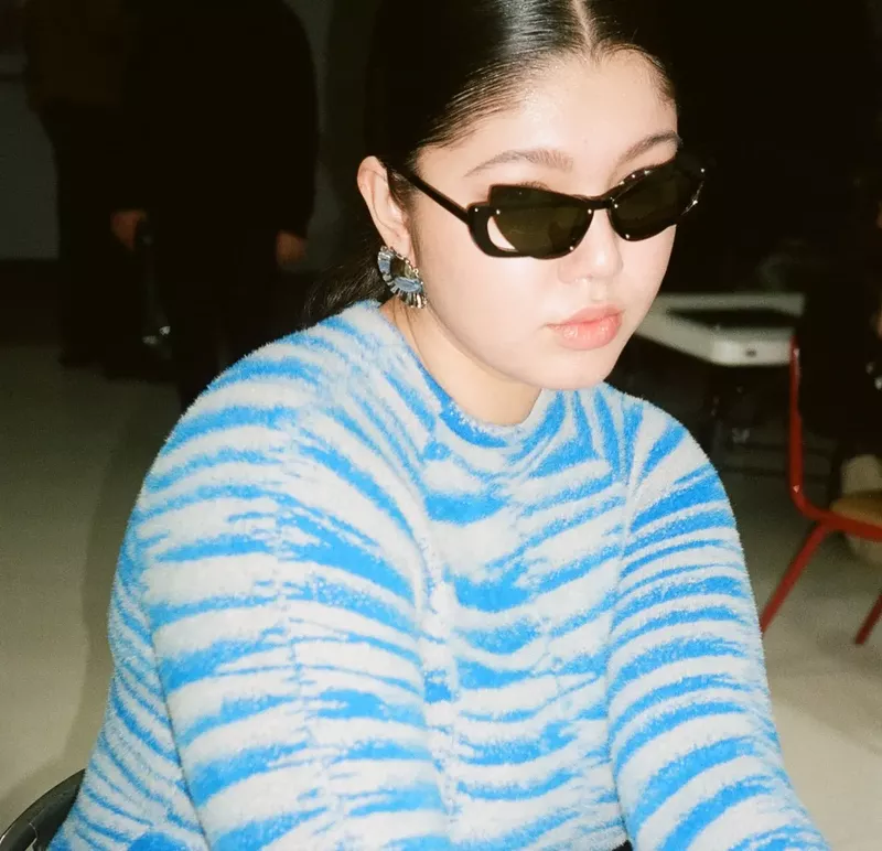 Portrait of a young woman with black sunglasses, earrings, and a blue-and-white striped sweater indoors in soft lighting.