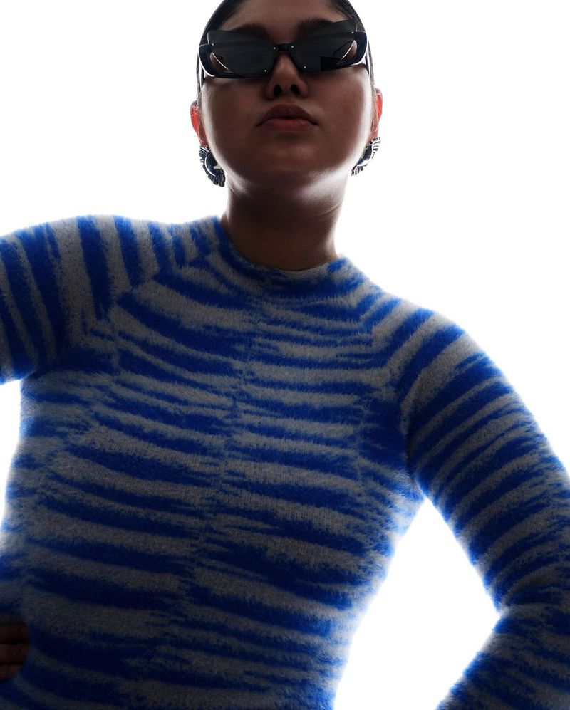 Stylish woman with sunglasses poses in a blue and white patterned sweater against a bright white background in a fashion shot.