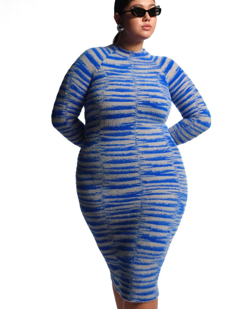 Adult woman poses in a bodycon sweater dress with gray and blue horizontal stripes against a white backdrop in a studio setting.