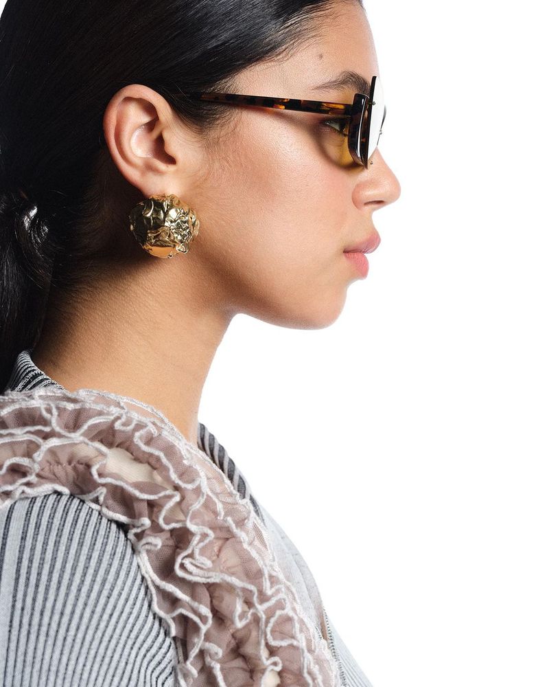 Profile portrait of a woman with tortoise-shell sunglasses and gold earrings in a striped collared shirt on a white background.