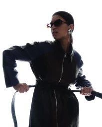 Model wearing dark sunglasses and a belted coat pulling a belt against a bright white seamless background
