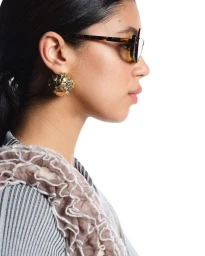 Profile portrait of a woman wearing tortoiseshell sunglasses and a gold statement earring against a white background.