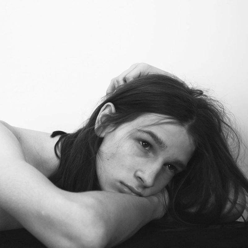 Black and white portrait of a young adult with long hair. The person is laying down with their arms crossed, gazing at the camera.