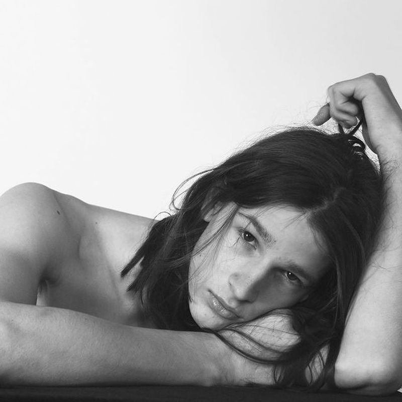 Black and white portrait of a topless young man with long dark hair, tear on face, resting on his arms against a white background.
