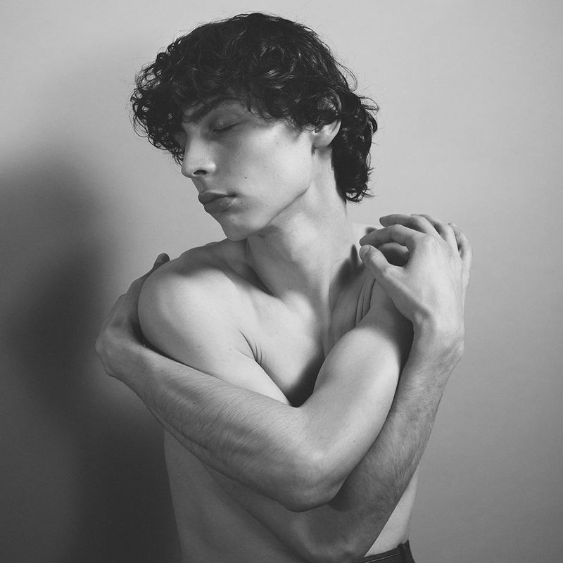 Black and white portrait of a young adult male embracing himself with closed eyes in a photo studio setting with a neutral background.