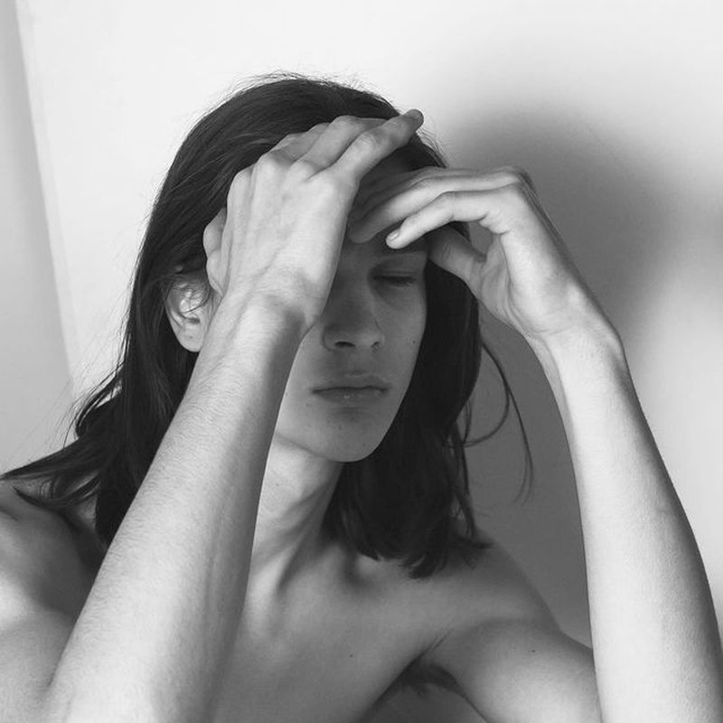 Black-and-white studio portrait of a bare-chested adult male with long hair, eyes closed, and hands touching his forehead in a thoughtful pose.