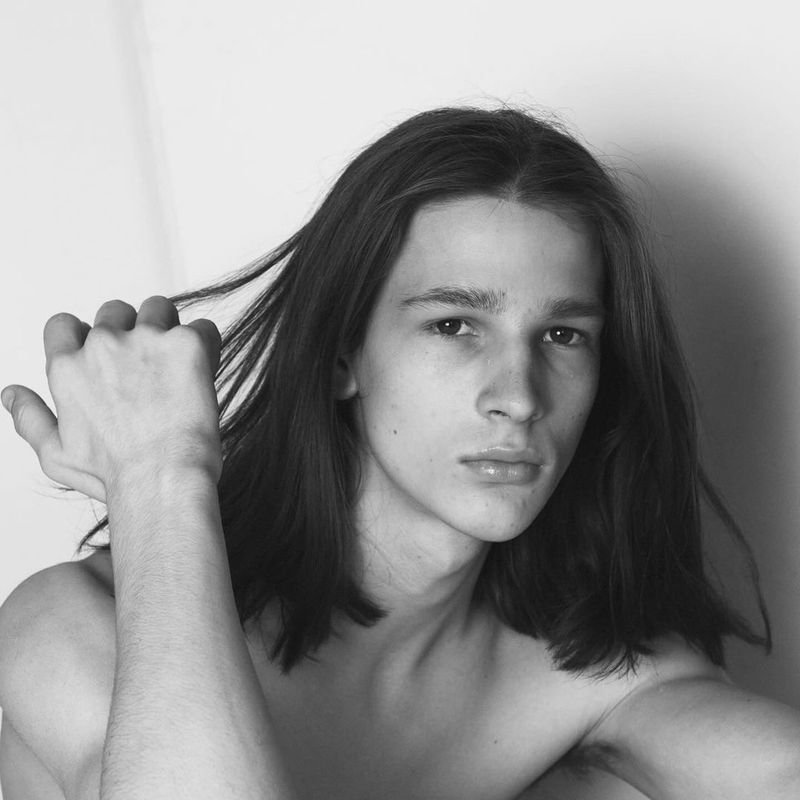 Portrait of a shirtless young adult male with long brown hair posing in front of a white background, holding his hair with his hand.