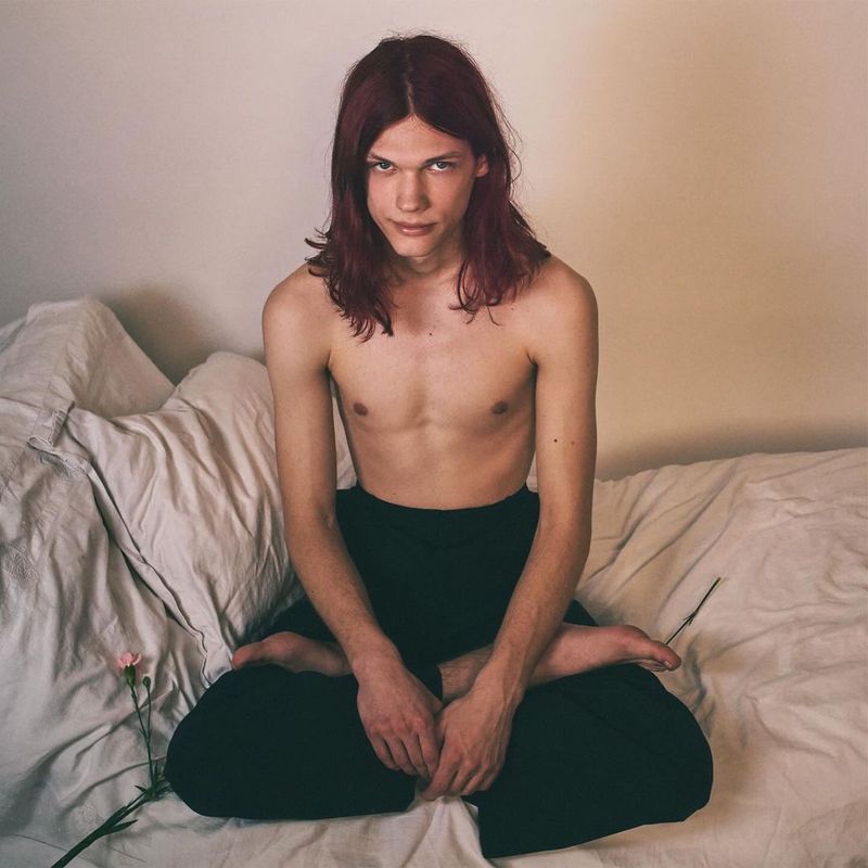 A topless person with long auburn hair sits cross-legged on a rumpled white bed in front of a white wall, with a pink carnation beside them.