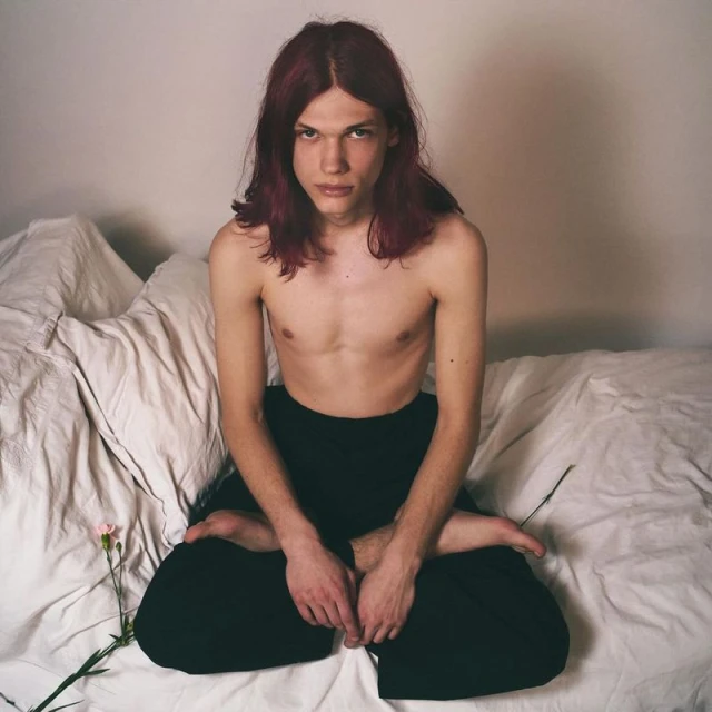 A shirtless person with long, dark red hair sits cross-legged on a white bed next to a pink flower in a brightly lit indoor setting.