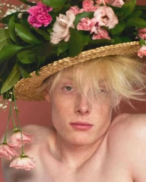 Close-up portrait of a fair-skinned model wearing a large straw hat adorned with fresh pink flowers, photographed by David Sims for Loewe.