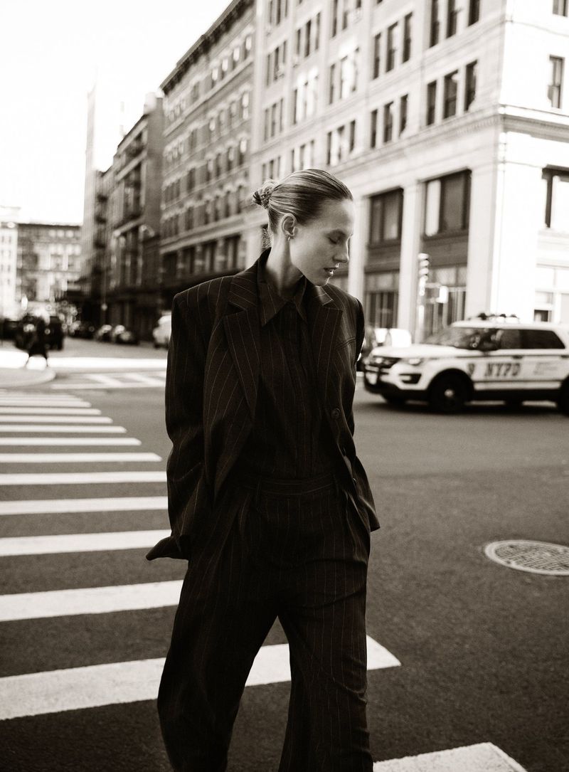 A woman in a pinstripe suit walks across a crosswalk in a city setting, with buildings and an NYPD car in the background. Sepia tone.
