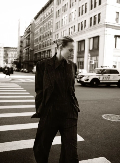 A woman in a pinstripe suit walks across a crosswalk in a city setting, with buildings and an NYPD car in the background. Sepia tone.