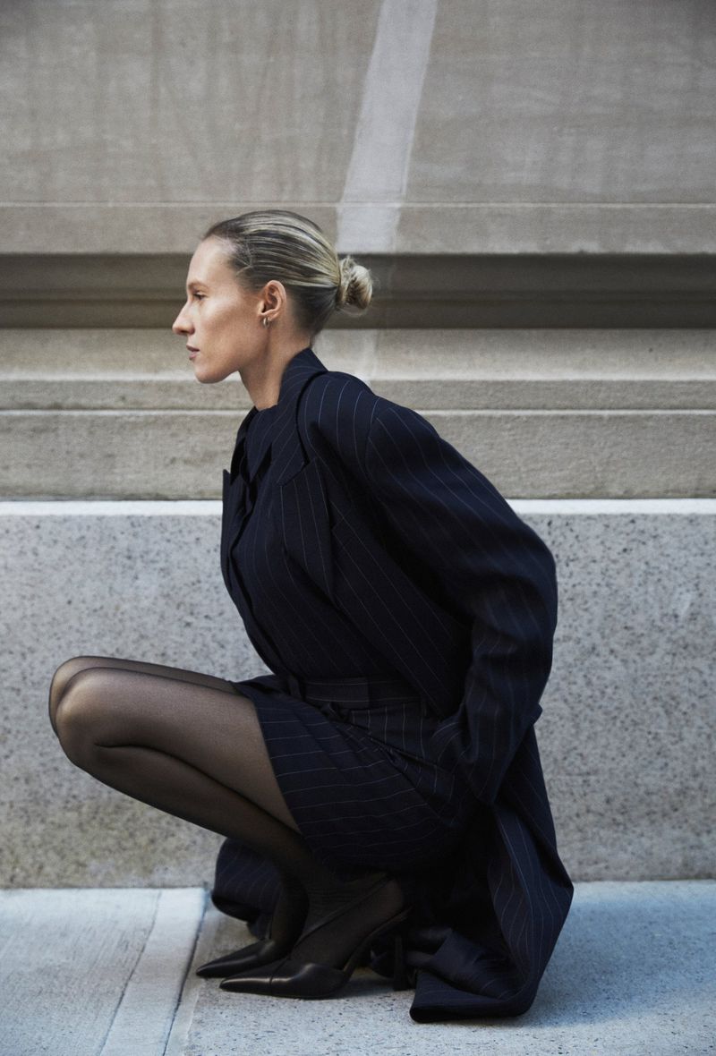Woman in a pinstripe suit and black tights crouching in an urban setting, looking off to the side, showcasing fashion and style.