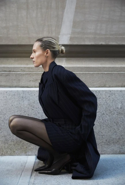 Woman in a pinstripe suit and black tights crouching in an urban setting, looking off to the side, showcasing fashion and style.