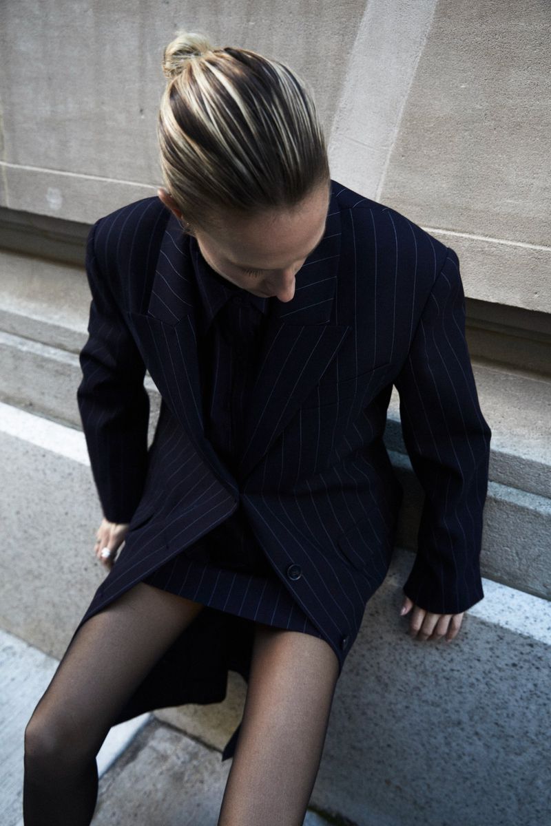 Blonde woman in pinstripe blazer and skirt, wearing black tights, sits on a concrete ledge, looking down, in a studio shot.