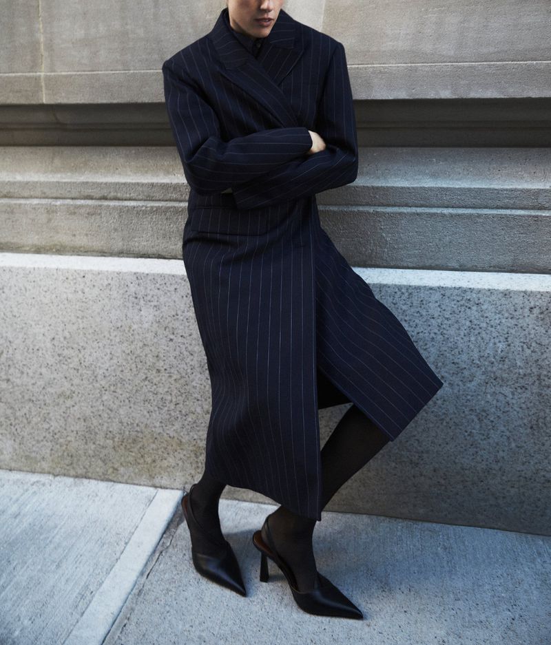 Woman in a navy blue pinstripe dress and black heels posing against a concrete wall in an urban environment, fashion photography.