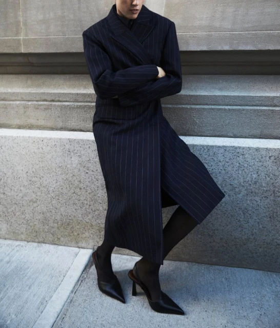 Woman in a navy blue pinstripe dress and black heels posing against a concrete wall in an urban environment, fashion photography.