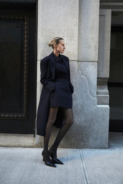 A woman poses in a dark pinstripe suit with matching skirt, top, and coat on a city street with a stone building behind her.
