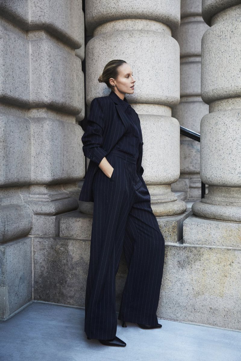 An adult female stands in front of an ornate building wearing a dark pin-striped suit with wide-leg pants in an urban environment.