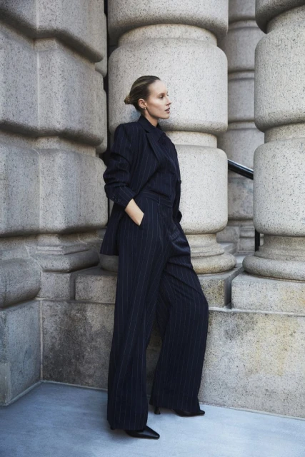 An adult female stands in front of an ornate building wearing a dark pin-striped suit with wide-leg pants in an urban environment.