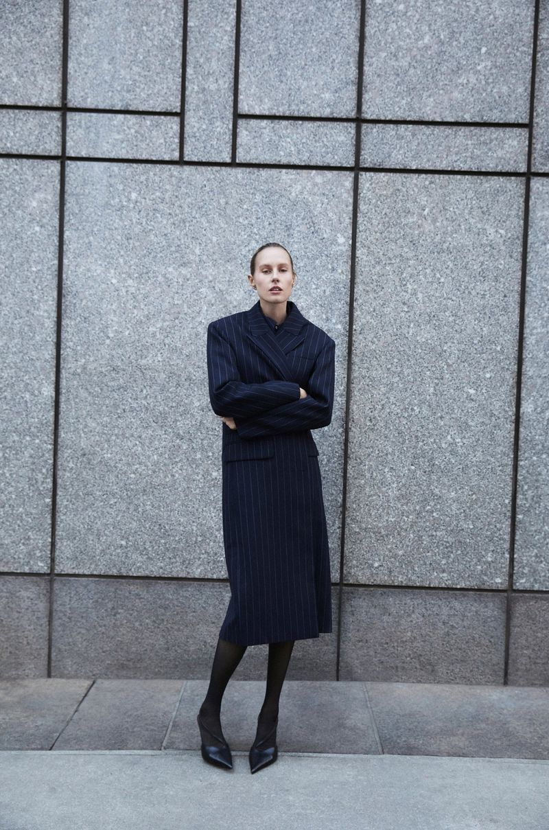 A woman in a pinstripe suit stands in front of a grey wall with geometric designs. She is wearing black tights and heels.