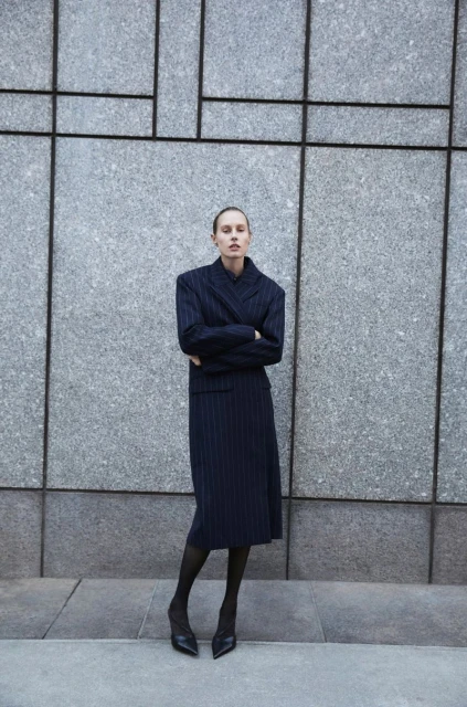 A woman in a pinstripe suit stands in front of a grey wall with geometric designs. She is wearing black tights and heels.