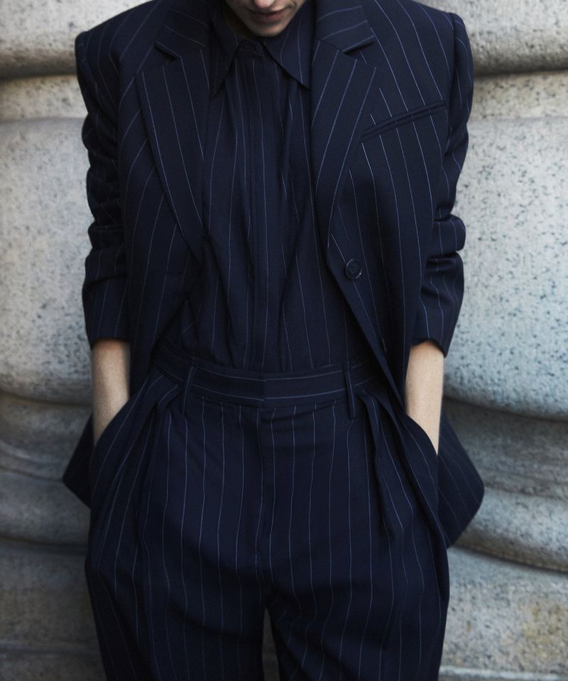 Model wears a dark-blue pinstripe suit with hands in pockets. The suit includes a jacket, collared shirt, and pants. The background is a neutral wall.