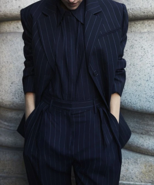 Model wears a dark-blue pinstripe suit with hands in pockets. The suit includes a jacket, collared shirt, and pants. The background is a neutral wall.