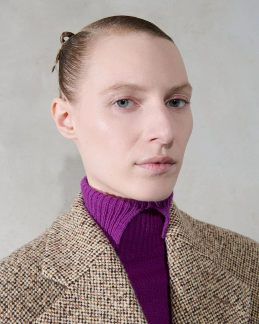 Headshot of a light-skinned young adult female model with hair in a bun, wearing a purple sweater and tweed jacket against a gray background.