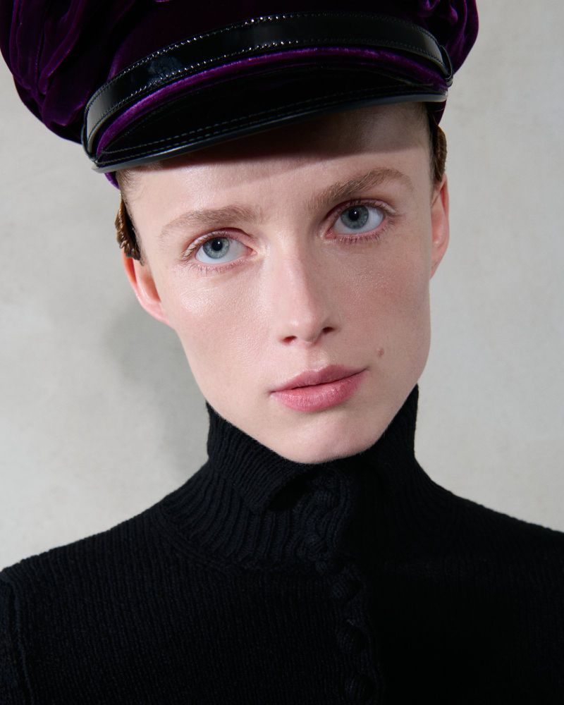 Close-up portrait of a woman with fair skin wearing a black turtleneck sweater and a purple velvet cap in a neutral indoor setting.