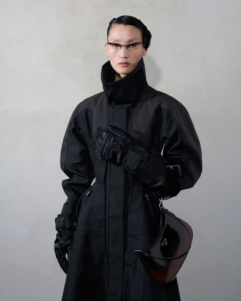 Asian woman in black coat and gloves, glasses, with helmet-like bag. Fashionable, edgy look against neutral background.