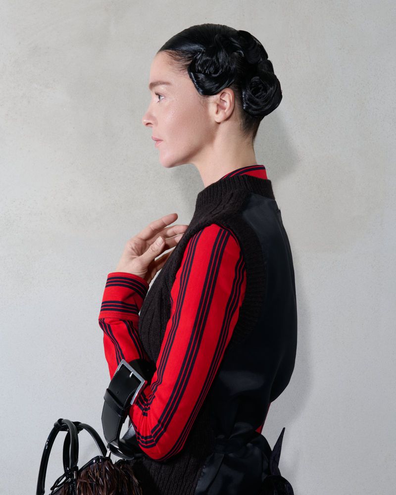 Profile view of a woman with sculpted black hair, wearing a red striped shirt and black vest against a neutral textured background.
