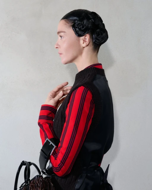 Profile view of a woman with sculpted black hair, wearing a red striped shirt and black vest against a neutral textured background.