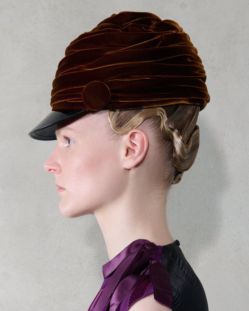 Woman with blonde hair styled up and wearing a brown velvet peaked hat with black brim, shot in a photo studio with light grey backdrop.