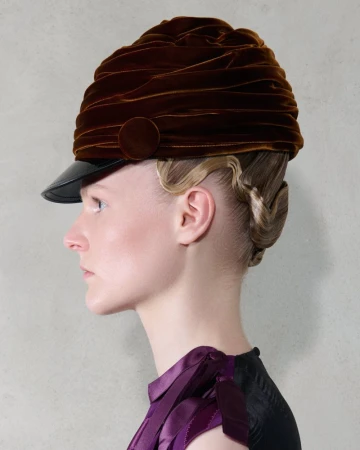 Woman with blonde hair styled up and wearing a brown velvet peaked hat with black brim, shot in a photo studio with light grey backdrop.