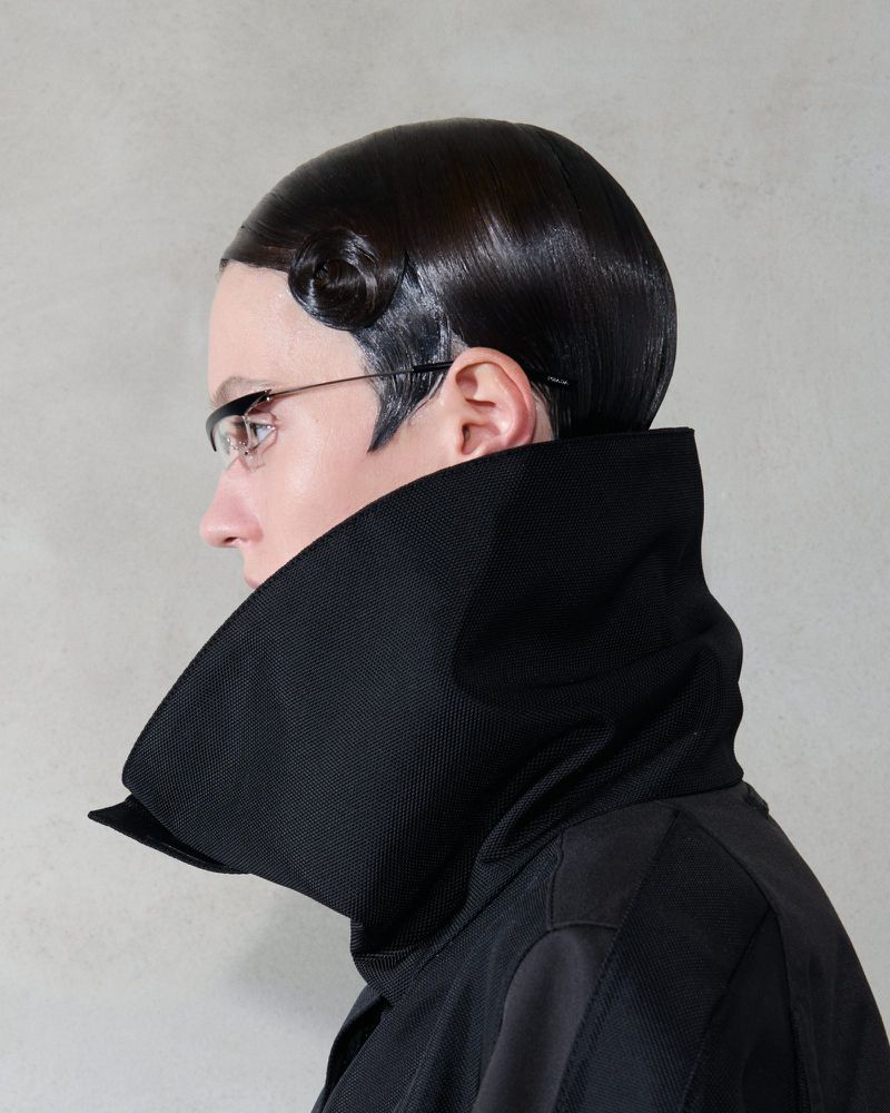 Woman in profile wearing glasses and a high-collared black jacket with retro hairstyle, standing indoors against a neutral background.
