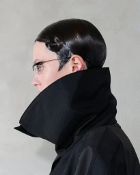 Profile portrait of a model with sculpted dark hair and high collar black coat against a neutral grey wall.